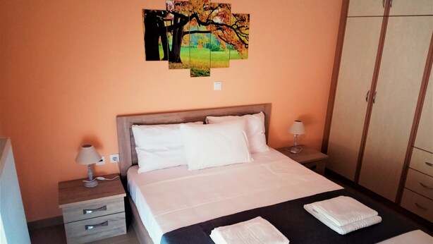 A neatly made bed with white linens, two bedside lamps, a nightstand with a lamp, a wooden headboard, and a wall-mounted painting of a tree. - Alkioni City Apartments (Chania)