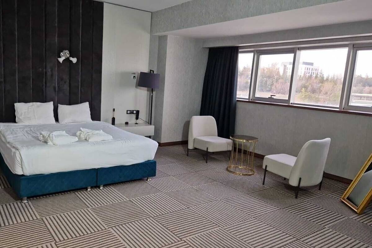 Ein Hotelzimmer mit einem großen Bett, zwei weißen Stühlen, einem kleinen runden Tisch und einem großen Fenster mit Blick auf Gebäude.