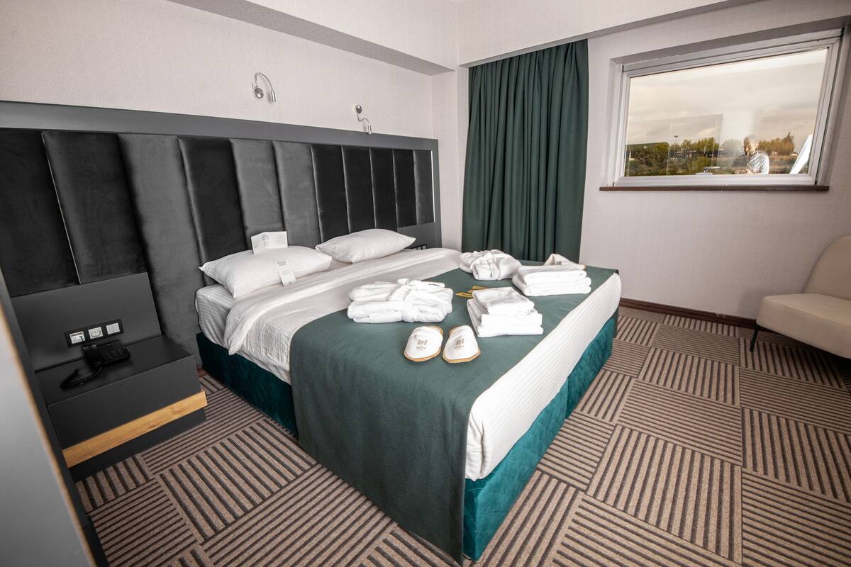 Ein Hotelzimmer mit einem großen Bett, einem Nachttisch mit Telefon und einem Fenster mit Vorhängen.