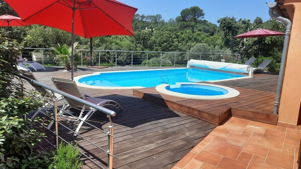 Une terrasse en bois avec une piscine, deux parasols rouges et une clôture en métal.