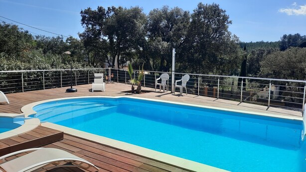 Piscina com deck de madeira, rodeada por guarda-corpo de metal e cadeiras de plástico brancas.