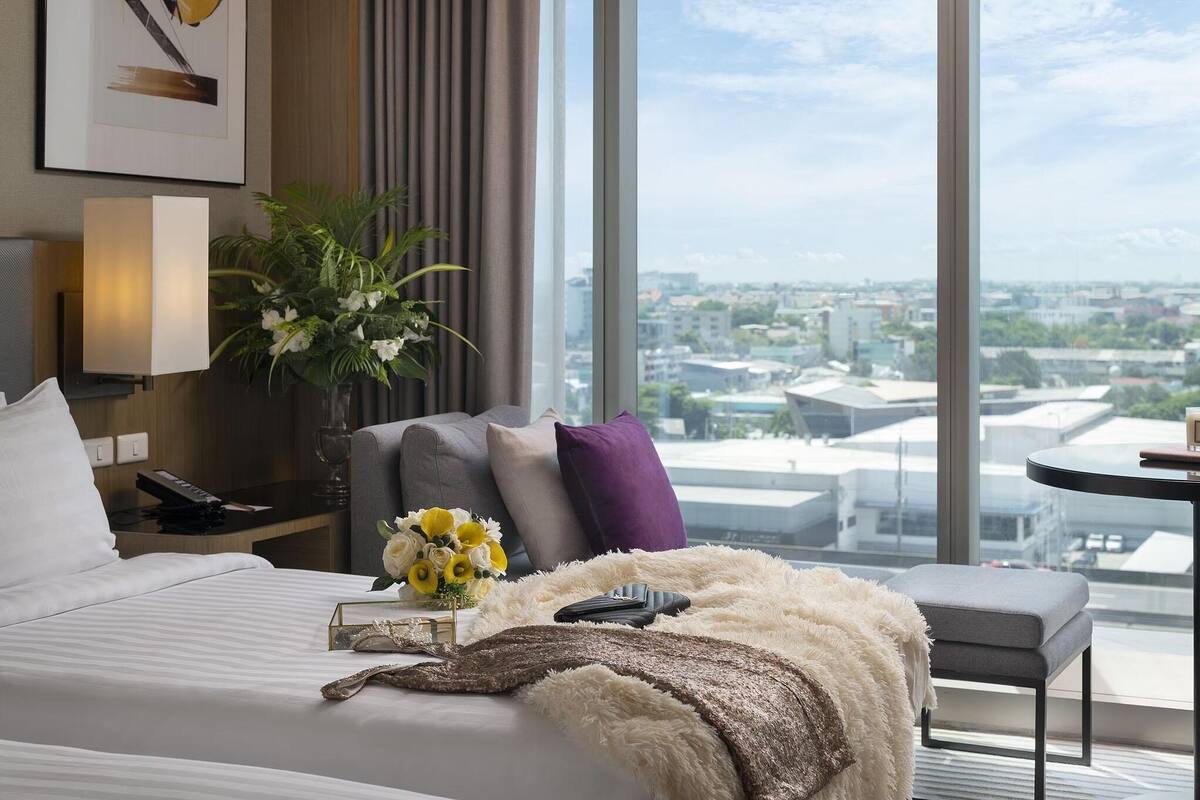 A hotel room with a large window, a bed with white bedding, a gray armchair, a small table, and a decorative plant.