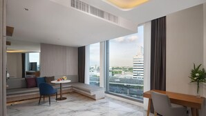A modern hotel room with a large window, a seating area with a round table, and a wooden desk. - Asawin Grand Convention Hotel (Bangkok)