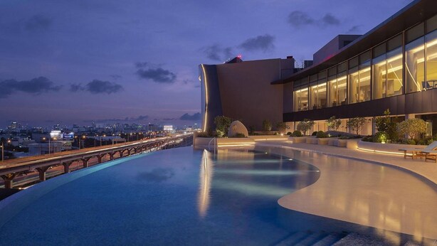 A modern building with a curved infinity pool, a cityscape view, and a bridge in the background. - Asawin Grand Convention Hotel (Bangkok)