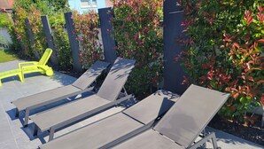 Zona patio con tre lettini prendisole e una sedia gialla, circondata da un recinto in legno e vegetazione.