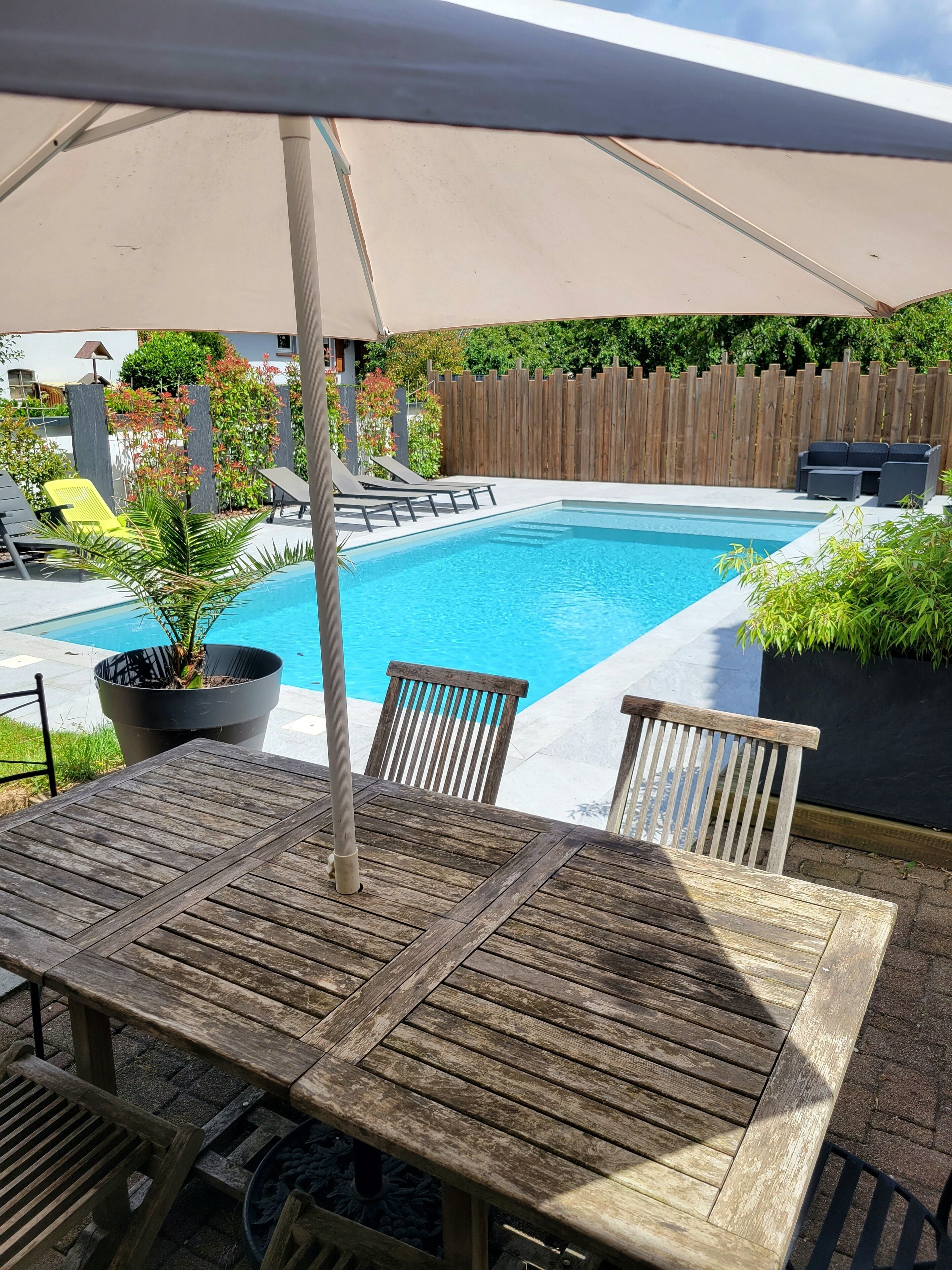 A pool area with a wooden table and chairs, surrounded by a wooden fence and greenery.