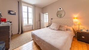 Ein Schlafzimmer mit einem großen Bett, einem Nachttisch mit Lampe, einem Schminktisch und einem Fenster mit Vorhängen.