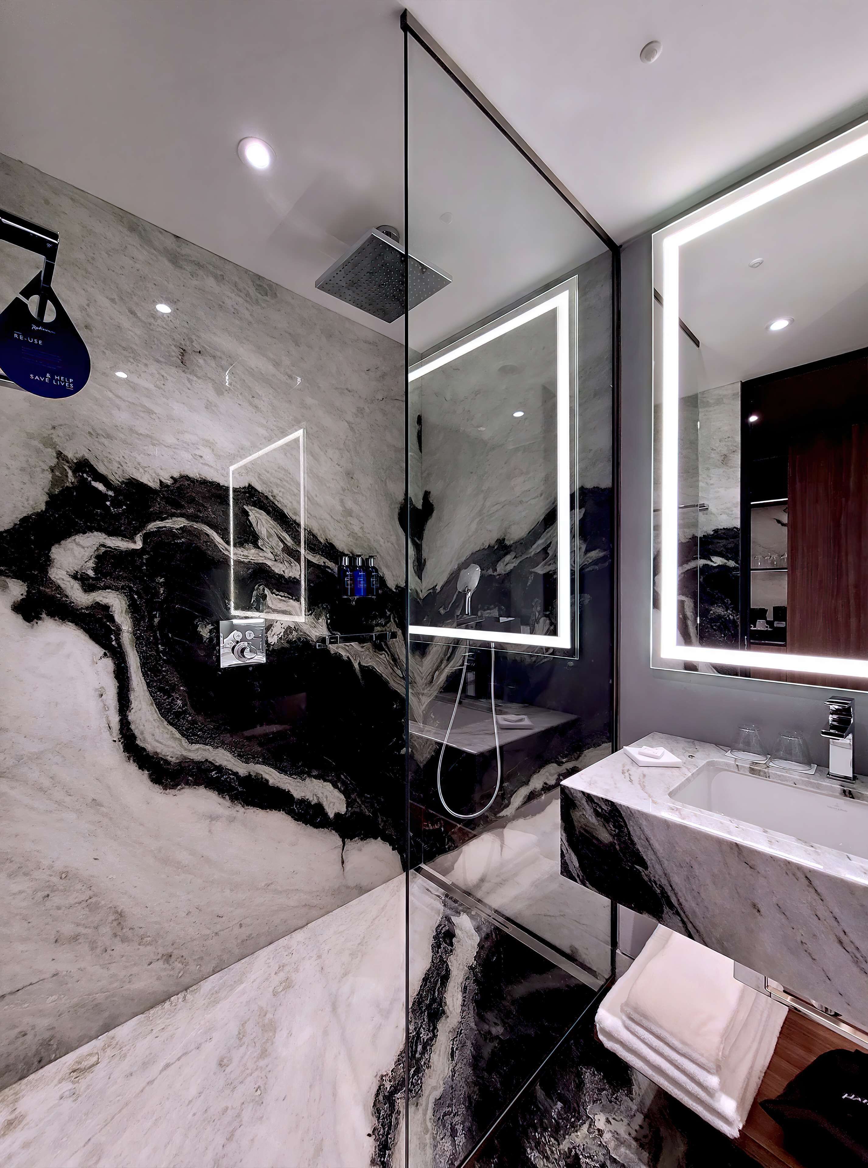 A modern bathroom with a marble wall, a glass-enclosed shower, a large mirror, and a vanity with a sink and towel rack.