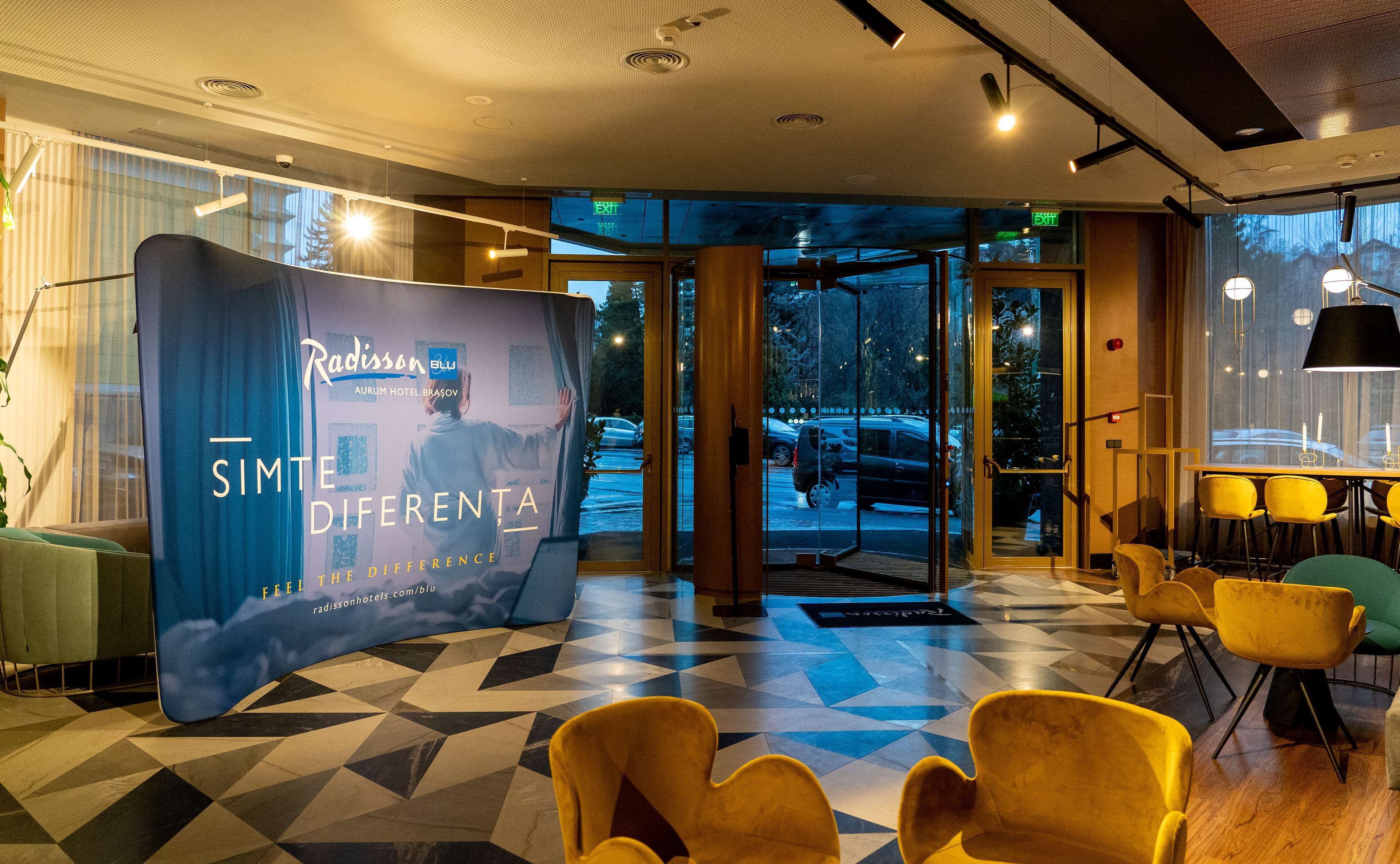 Un hall d’hôtel avec un sol à motifs géométriques, un présentoir publicitaire bleu et des chaises jaunes.