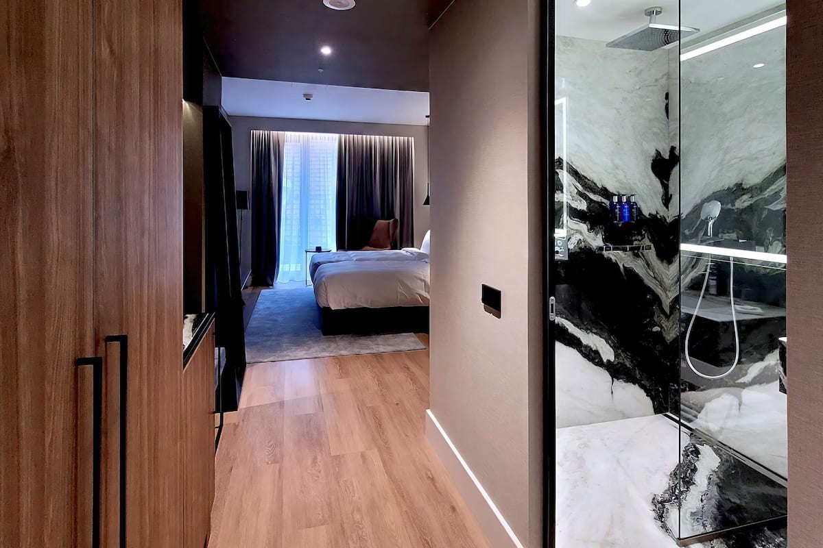 A modern hotel hallway with a wooden floor, a bed with a white headboard, and a glass shower area with marble tiles.