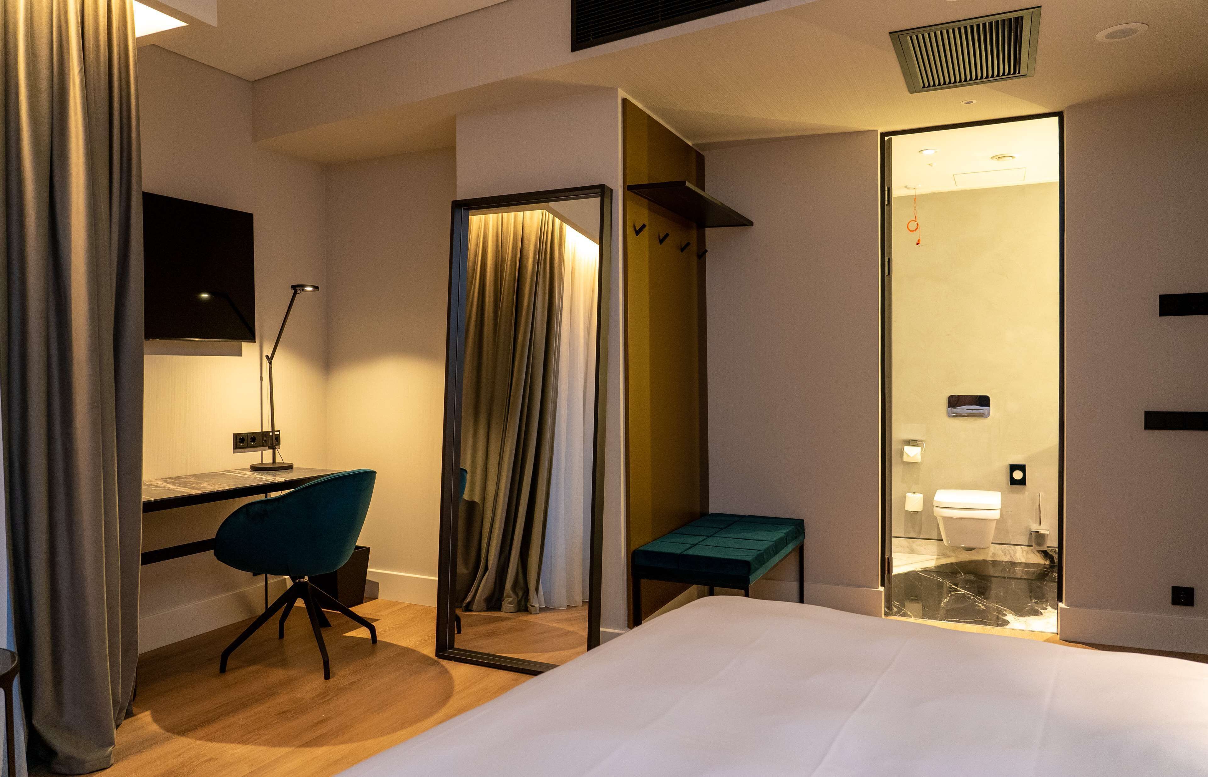 A hotel room with a bed, a desk with a chair, a mirror, a bench, and a bathroom visible through an open door.