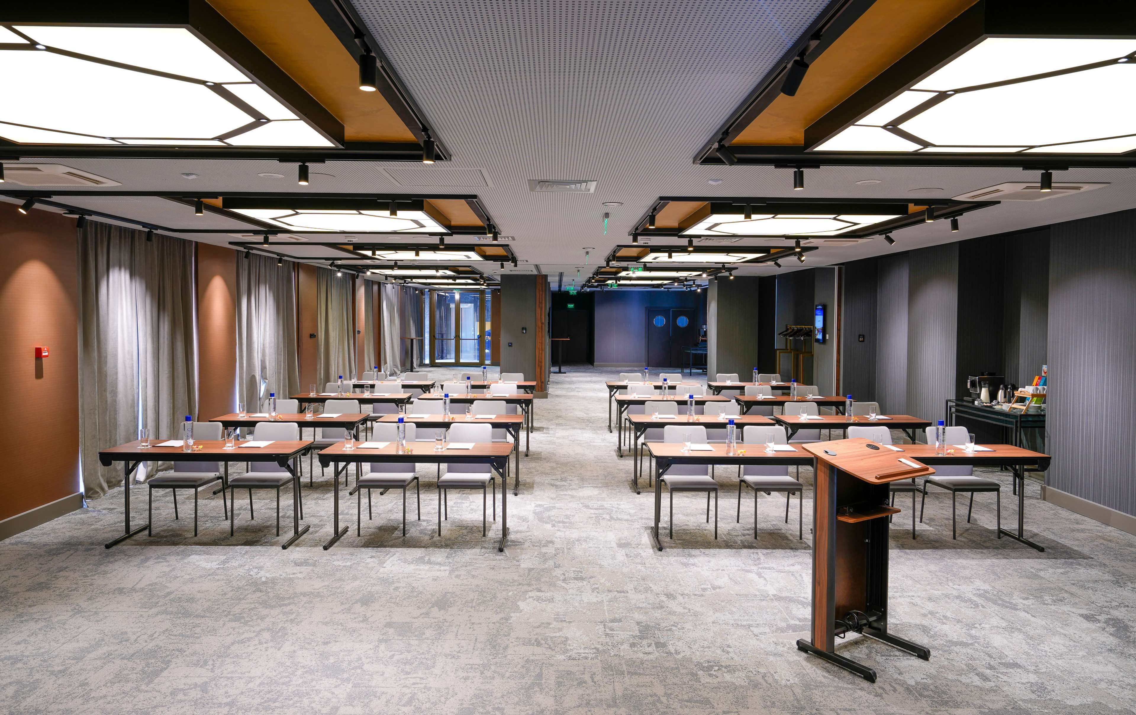A conference room with rows of tables and chairs, a podium, and a large window at the far end.