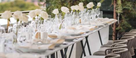 Una larga mesa de comedor adornada con flores blancas y cristalería, rodeada de sillas de mimbre.