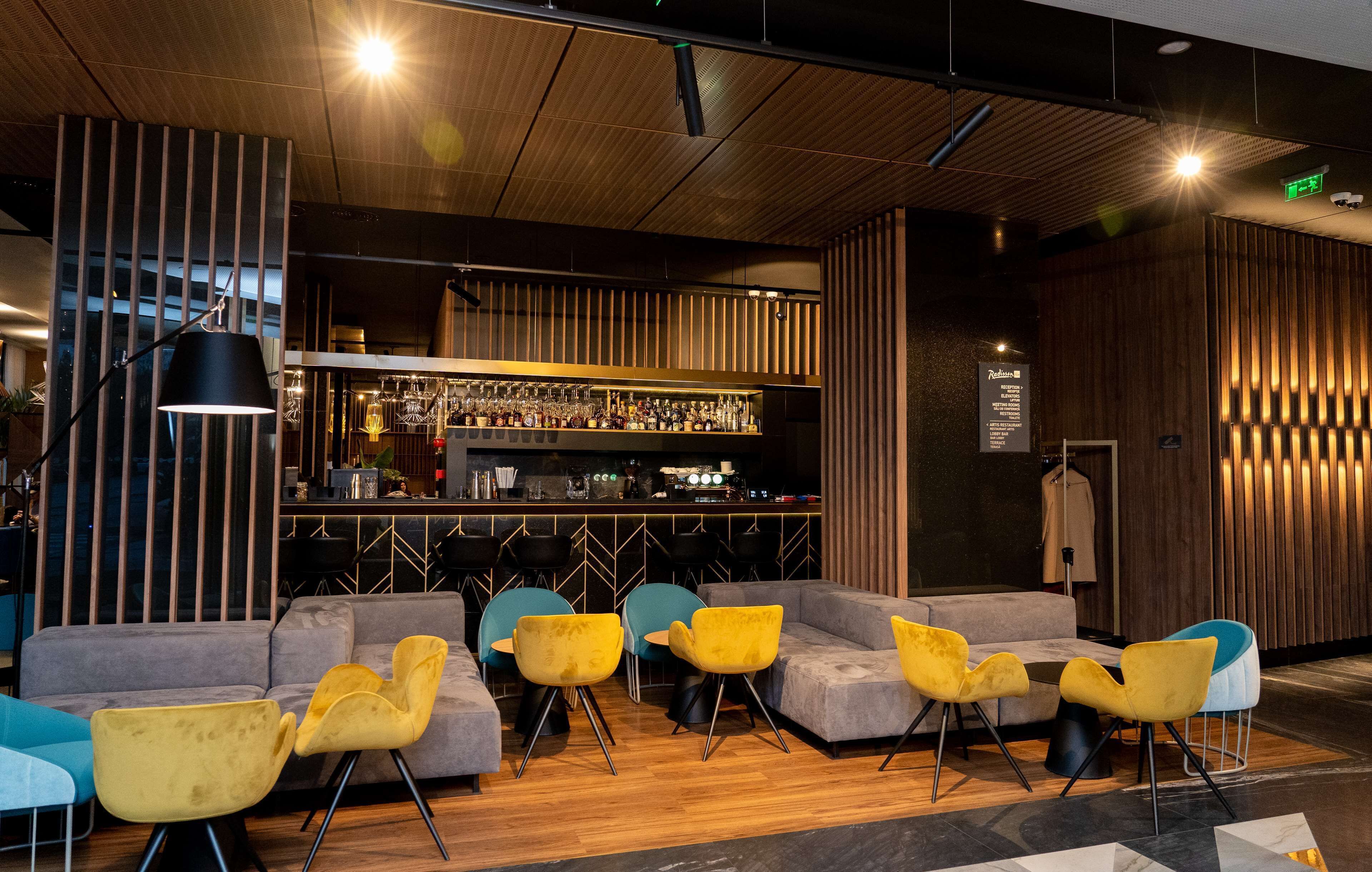 A modern bar area with a wooden ceiling, a bar counter with various bottles, and seating with yellow and blue chairs.