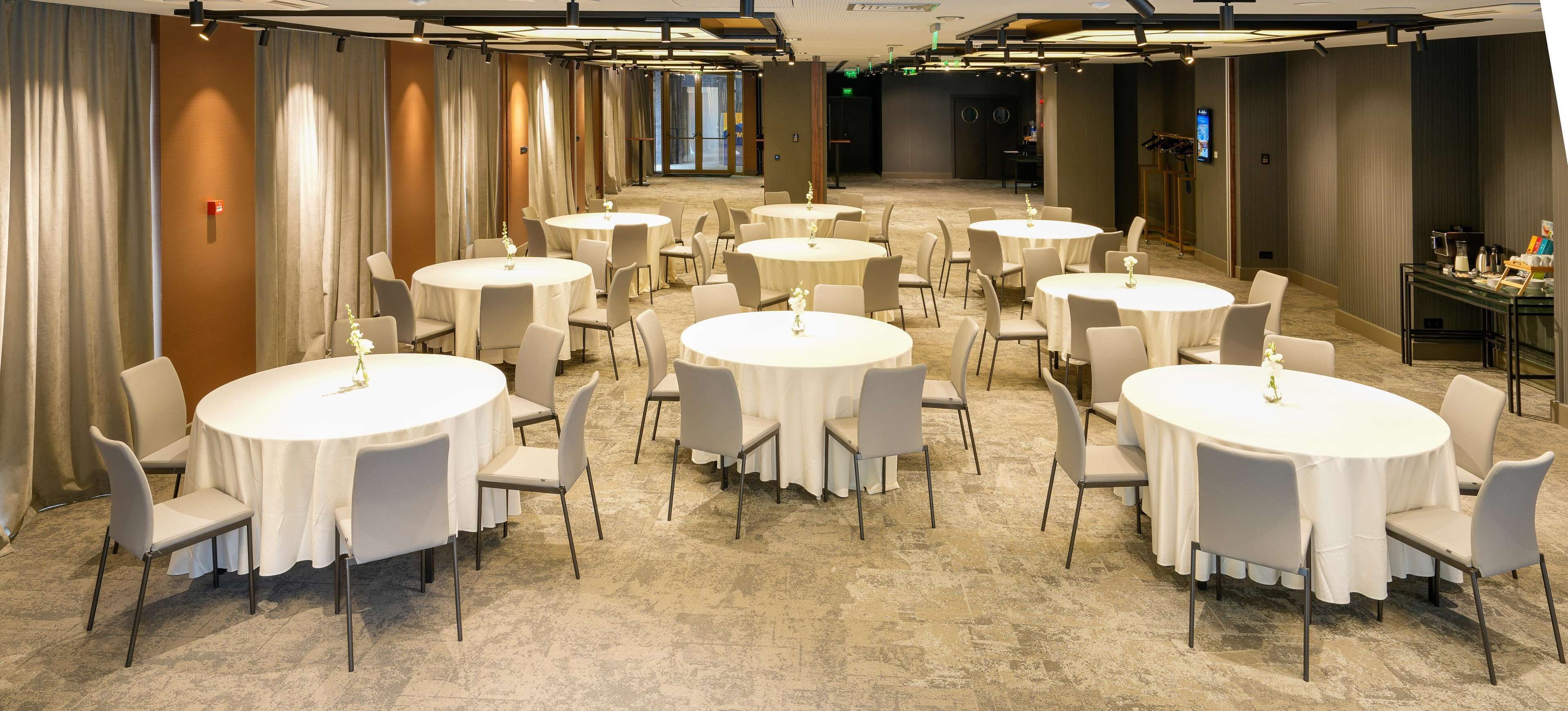 A hotel conference room with multiple round tables and chairs, carpeted flooring, and curtains along the walls.