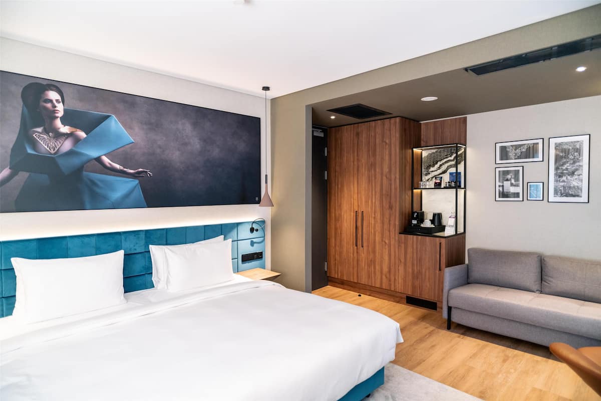 A modern hotel room with a large bed, a blue headboard, a grey sofa, and a wooden wardrobe.