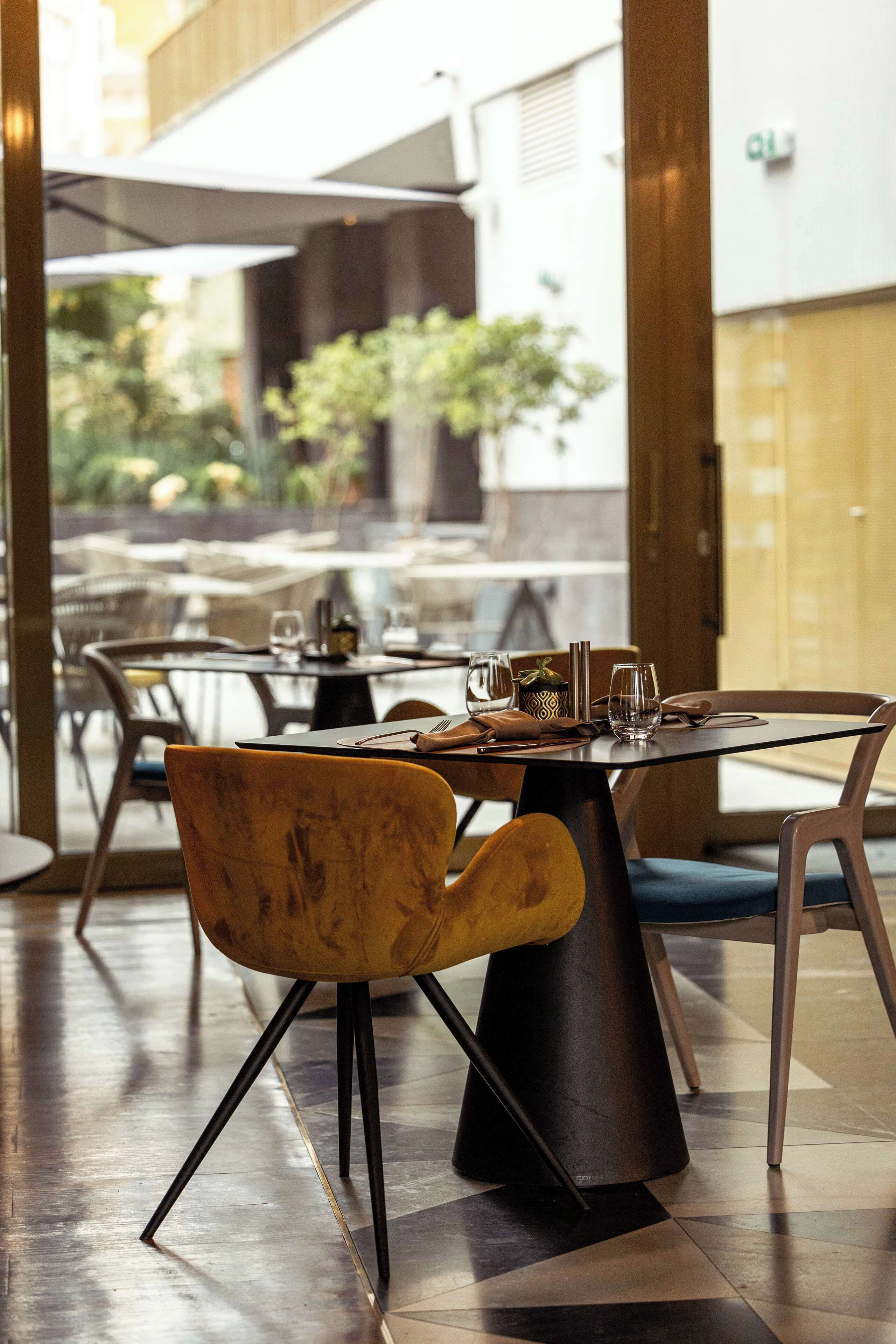 Interior de un restaurante con diseño moderno, con una mezcla de mobiliario de madera y metal, una zona de comedor de vidrio y vistas al área de mesas al aire libre.