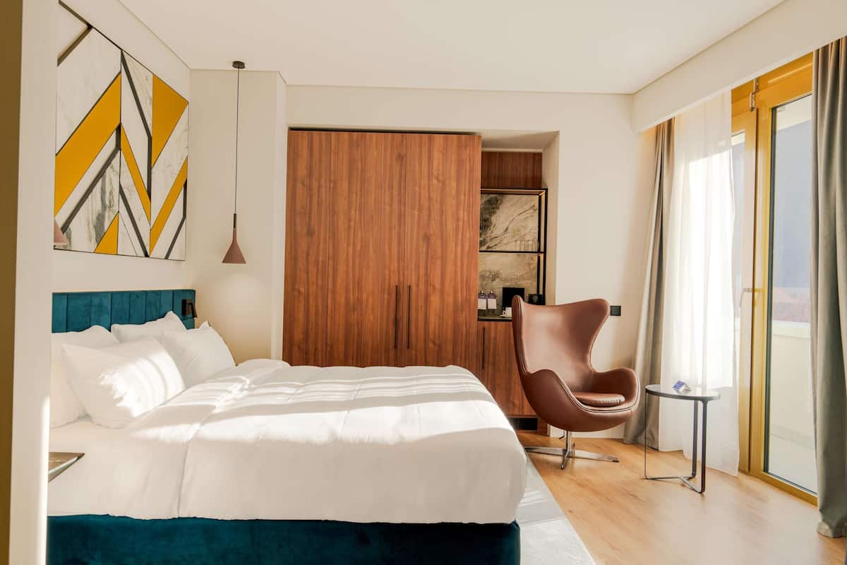 A modern hotel room with a large bed, a wooden wardrobe, a brown chair, and a view of the outdoors.