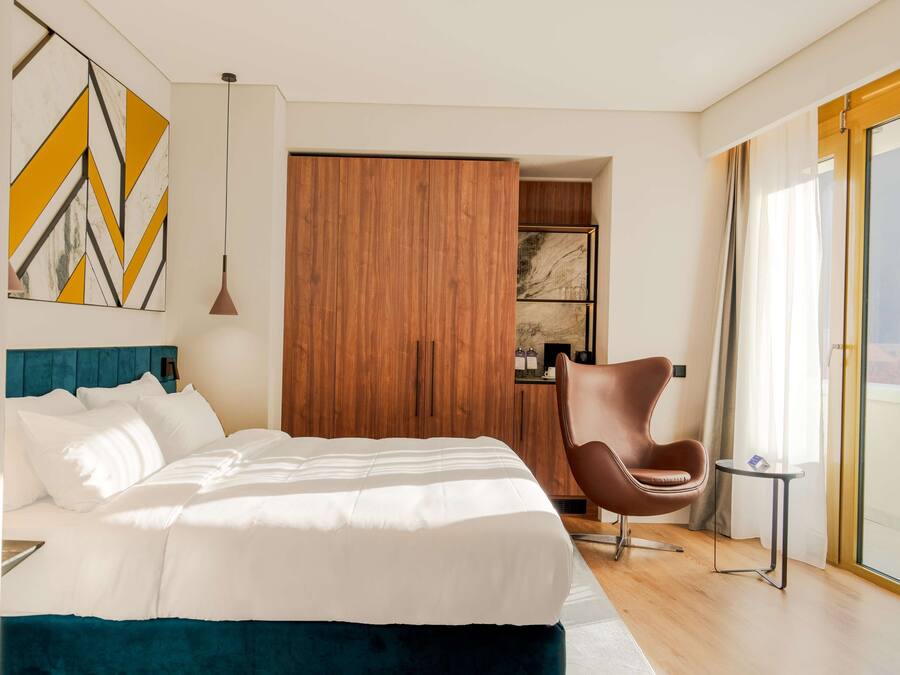 A modern hotel room with a large bed, a wooden wardrobe, a brown chair, and a view of the outdoors.