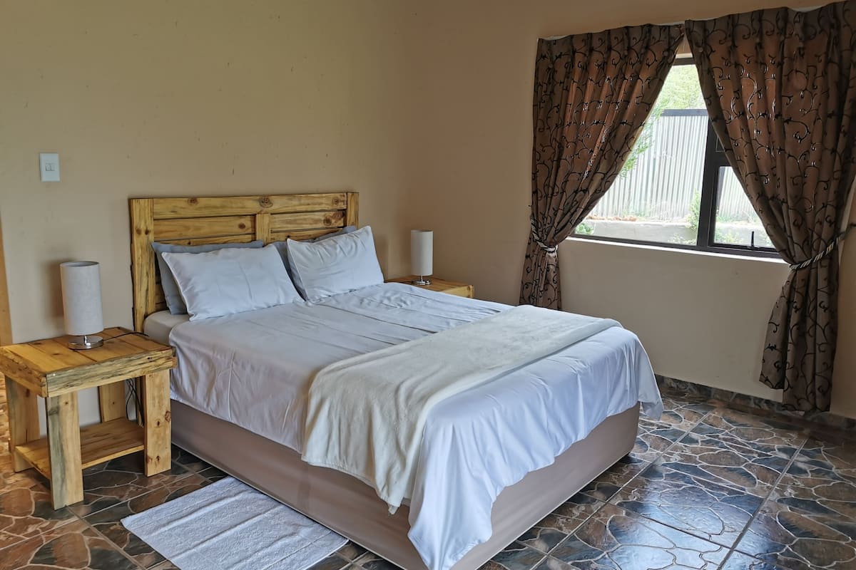 A bedroom with a wooden headboard, white bedding, two bedside tables with lamps, a window with curtains, and a stone-patterned floor.