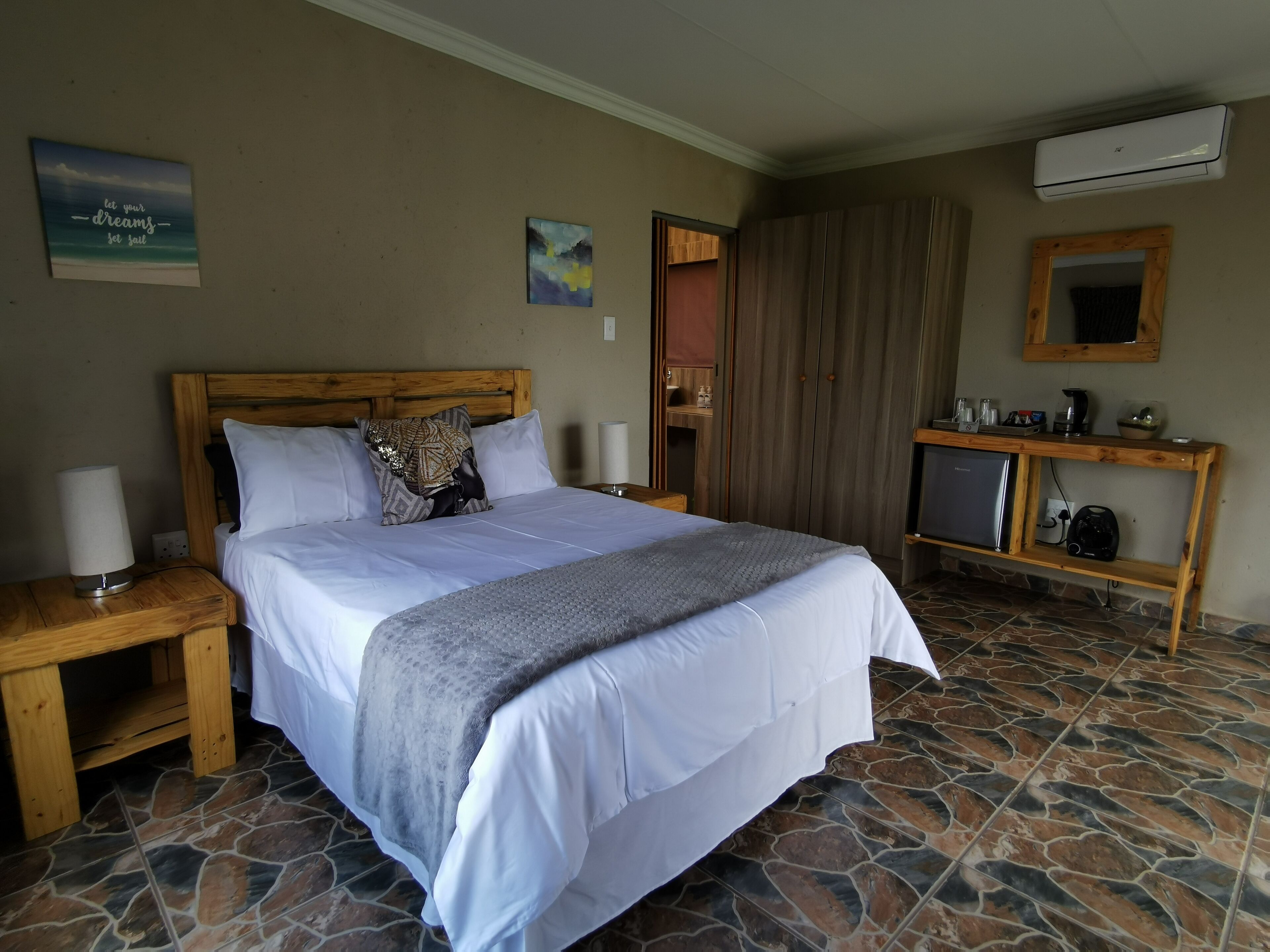 Une chambre à coucher bien aménagée, avec une tête de lit en bois, un lit recouvert de draps blancs, un plaid gris, une table de chevet avec une lampe, une petite étagère avec des articles de toilette et un sol carrelé en pierre.