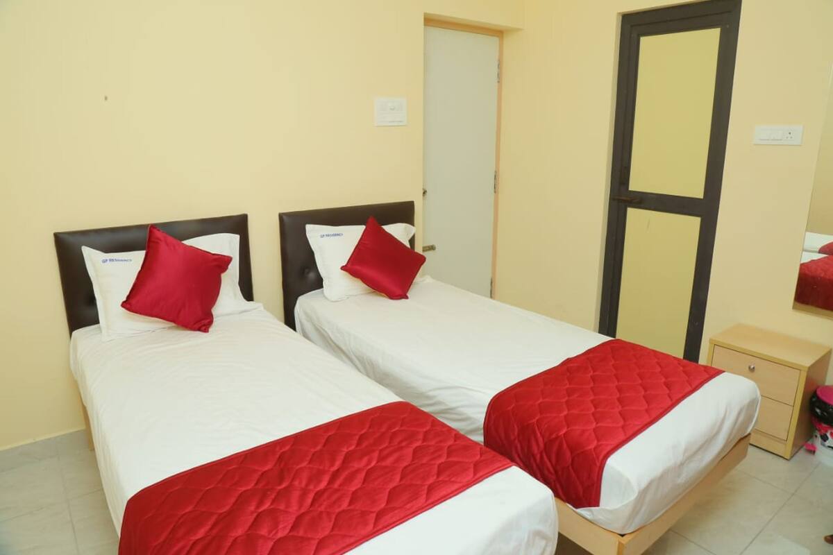 A hotel room with two beds, each with a red quilted bedspread and white linens. There is a wooden nightstand with a drawer, a small table, and a mirror on the wall.