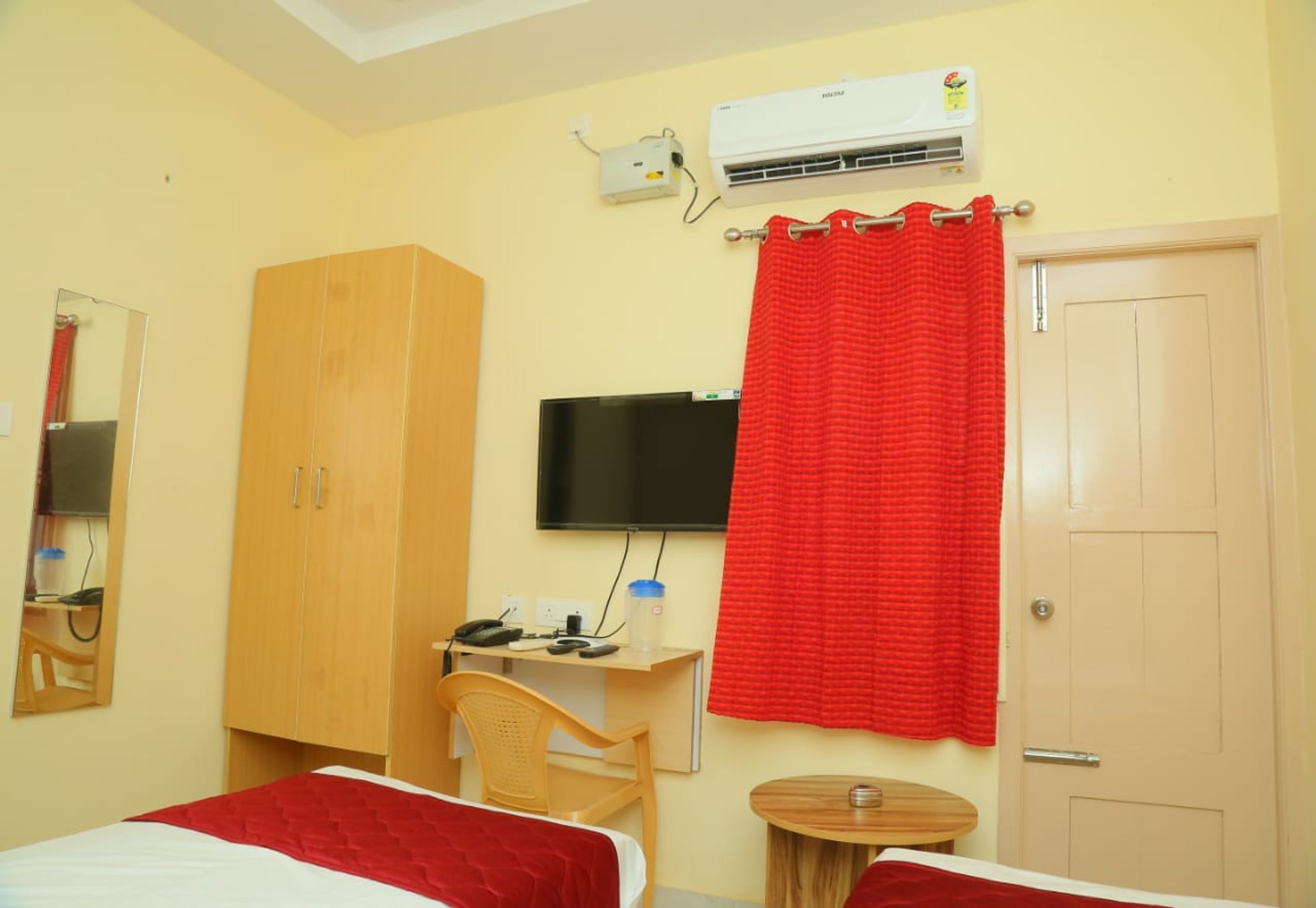 A hotel room with two beds, a desk with a chair, a television, an air conditioning unit, and a red curtain.