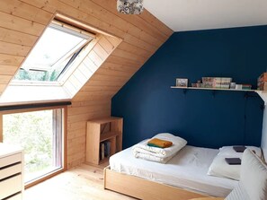 A bedroom with a sloped ceiling, a skylight, a bed with neatly folded blankets, a bedside table, and a shelf with books and decorative items. - 15 mn from the beaches, quiet and warm wooden house (Ploemel)