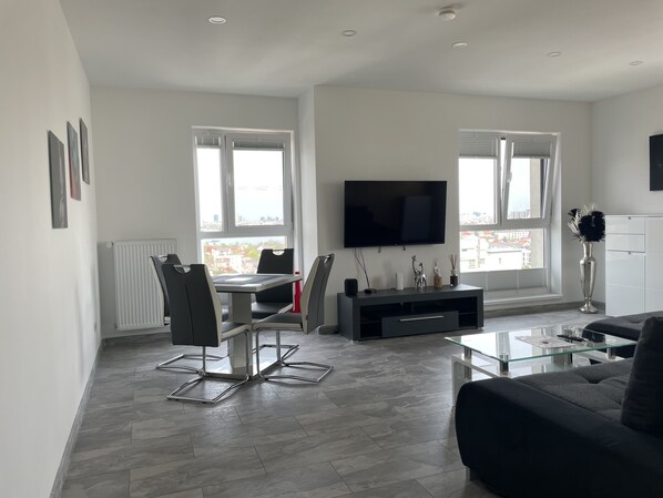 A modern living room with a dining area, a flat-screen TV, and a large window providing a city view.