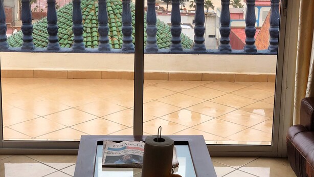 A room with a large window overlooking a beach, a glass-top table with a newspaper, and a leather couch.