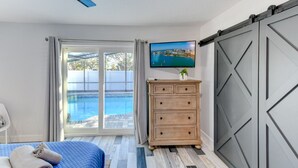 Un dormitorio con cama, cómoda, televisión y puerta corredera que da a una piscina.