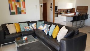 Uma sala de estar moderna com um sofá seccional, uma mesa de centro de vidro e uma área de cozinha ao fundo.