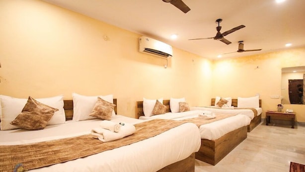 A hotel room with three beds, each with white bedding and brown pillows. There are ceiling fans, an air conditioning unit, and a small bedside table with a lamp. - Hotel Babua Hyness (Haridwar)