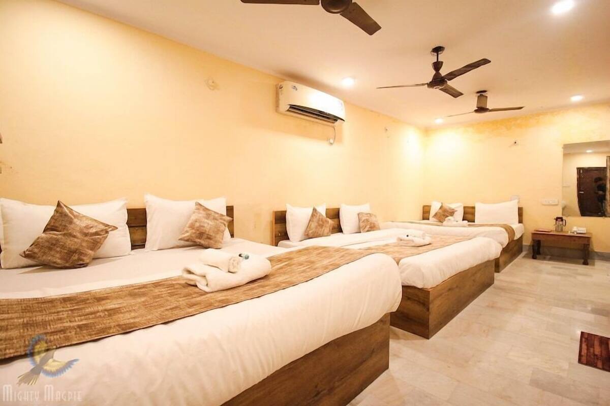 A hotel room with three beds, each with white bedding and brown pillows. There are ceiling fans, an air conditioning unit, and a small bedside table with a lamp.