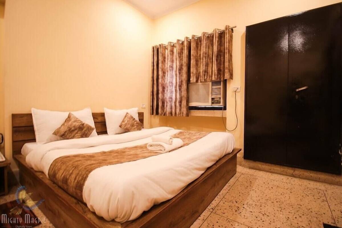 A double bed with white and brown bedding, a wooden headboard, a black wardrobe, and a small window with curtains.