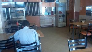 A restaurant interior with a person seated at a table, a television mounted on the wall, and a refrigerator in the background. - Pivot Hotel (Mwingi)