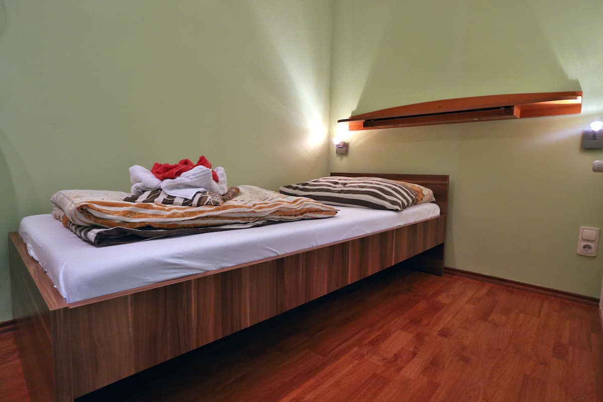 A single bed with a wooden frame, a white mattress, and striped bedding. There is a wall-mounted light above the bed and a power outlet on the wall to the right.