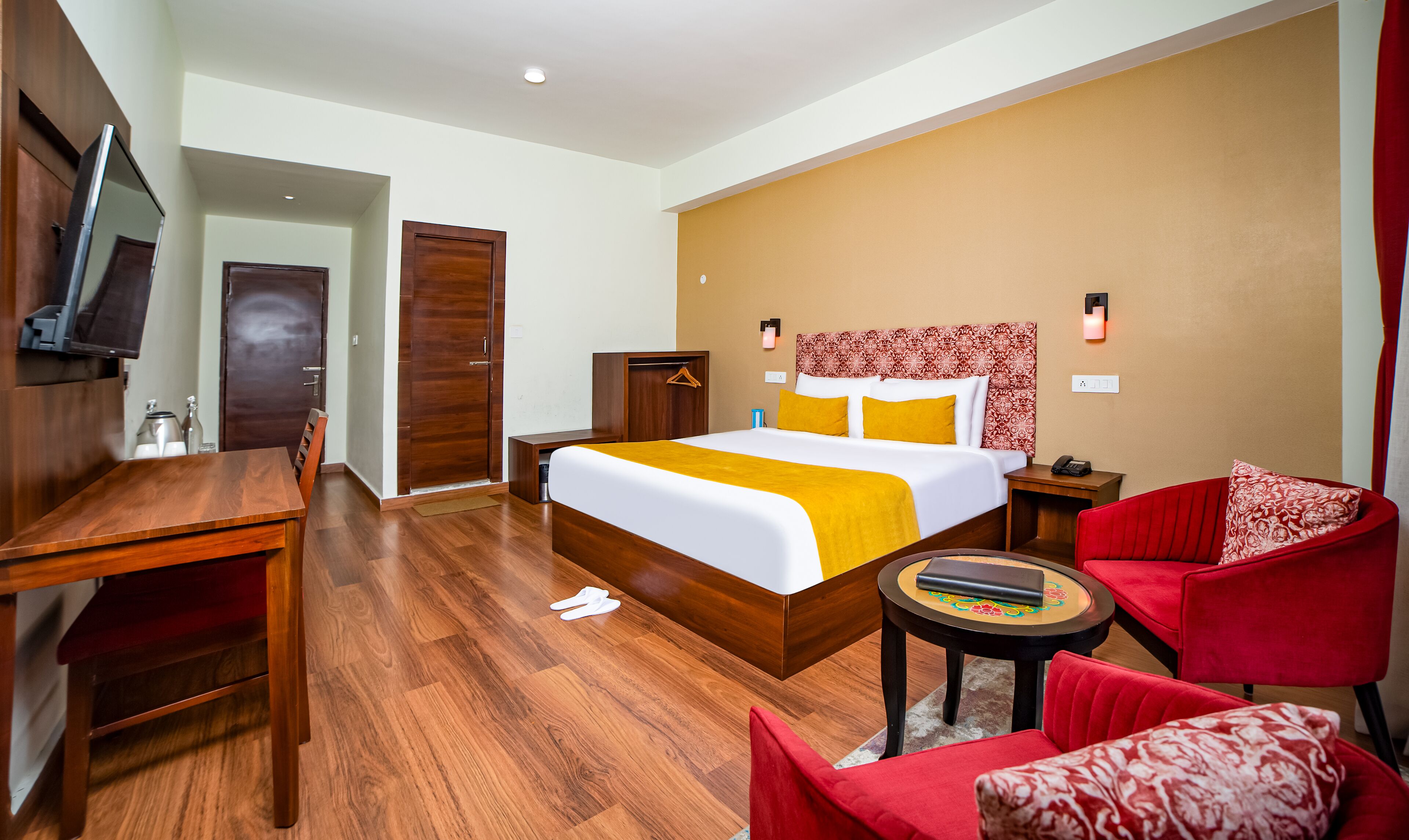Habitación de hotel con suelo de madera, cama con cubrecama blanco y amarillo, sillón rojo, una pequeña mesa redonda y un escritorio con silla.