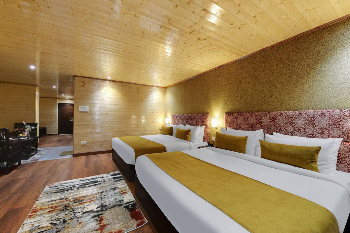 Habitación de hotel con techos de madera, dos camas con ropa blanca y almohadones color mostaza, una alfombra estampada y un pequeño escritorio con una silla.