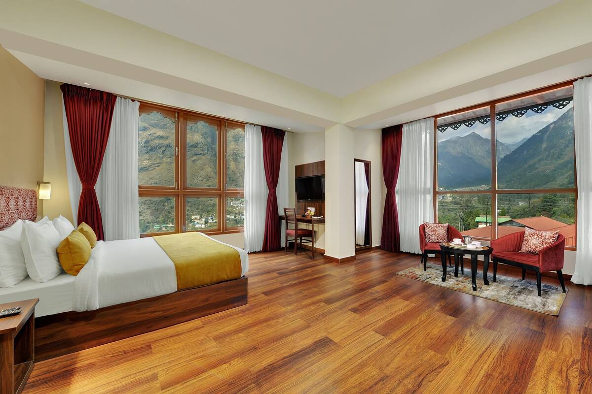 Habitación de hotel con cama, escritorio, silla y sofá. Amplias ventanas con cortinas y vistas a montañas.