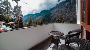 Un balcón con sillas de mimbre y una mesita, con vistas a un paisaje montañoso que incluye un valle y un pueblo lejano.