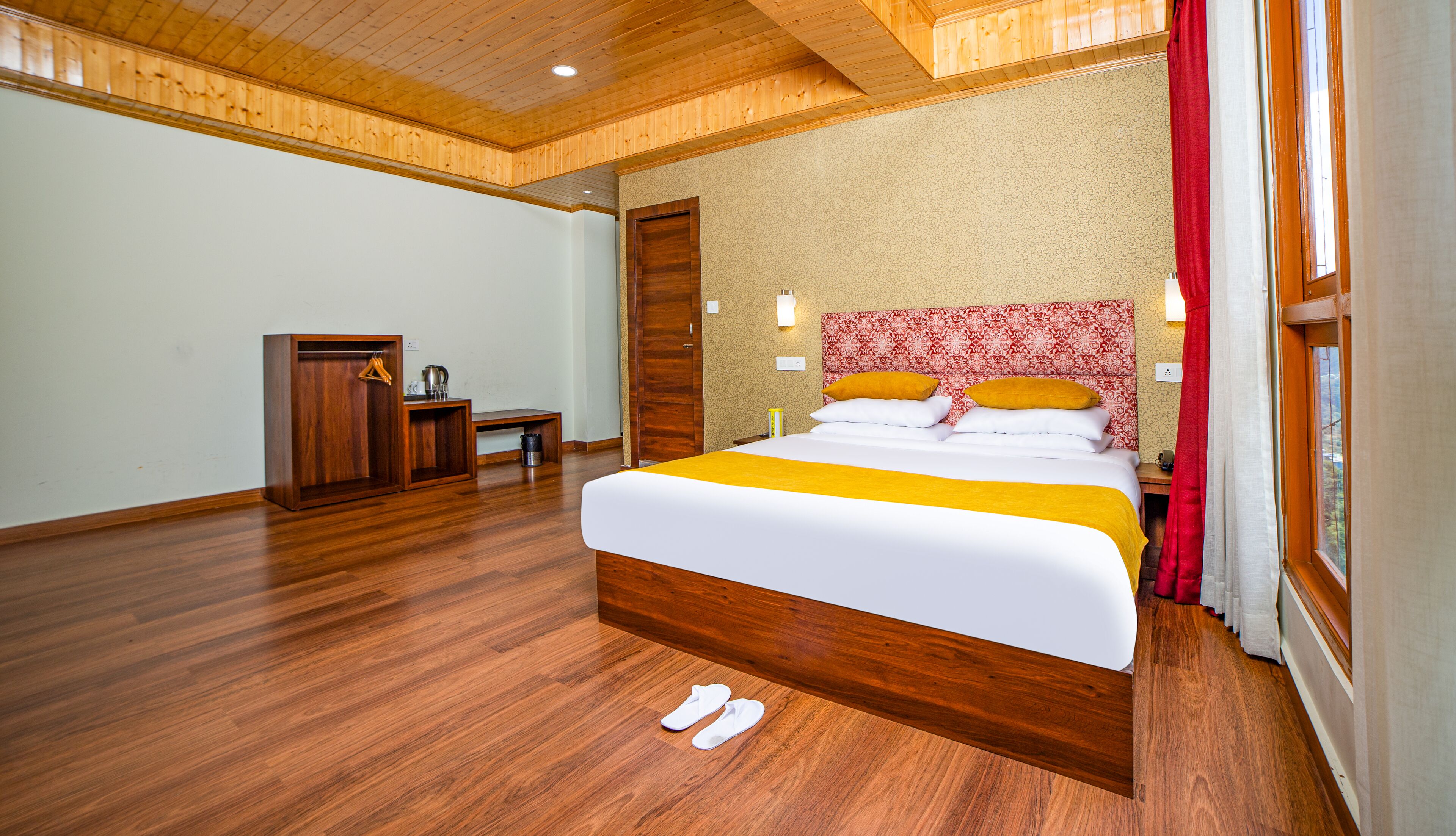 Een hotelkamer met een houten vloer, een bed met wit-geel beddegoed, een hoofdbord met een rood patroon, een houten garderobe en een klein bureau met een stoel.