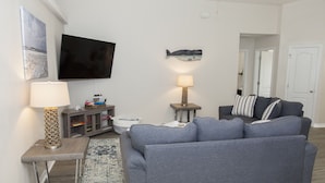 A living room with a grey sofa, a flat-screen TV mounted on the wall, a side table with a lamp, and a small cabinet. - Welcome to Beachy Bliss – Your Emerald Waters Escape! (Navarre)