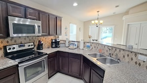 A kitchen with dark wood cabinets, a stainless steel microwave, a double oven, granite countertops, a double sink, and a chandelier. - Welcome to Beachy Bliss – Your Emerald Waters Escape! (Navarre)