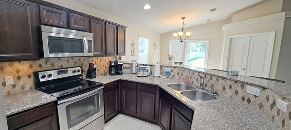 A kitchen with dark wood cabinets, stainless steel appliances, a granite countertop, and a window with blinds. - Welcome to Beachy Bliss – Your Emerald Waters Escape! (Navarre)