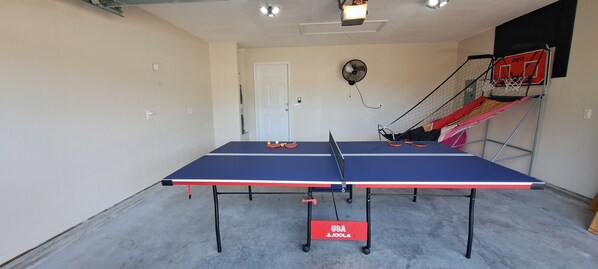 A ping pong table in a room with a door, a fan, and a basketball hoop.