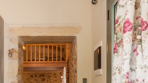 A narrow hallway leading to a bedroom with a stone wall, a wooden ceiling, a bed, and a nightstand with a lamp.