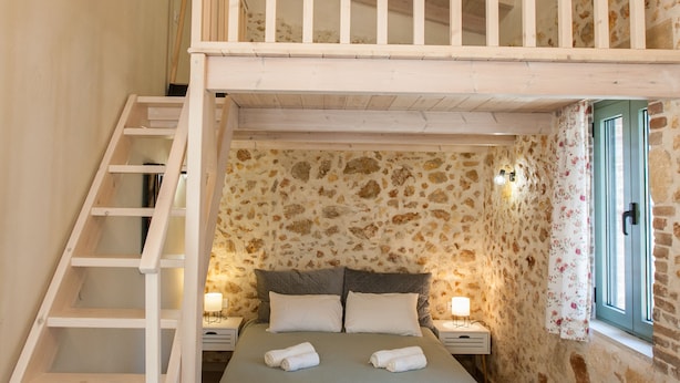 A bedroom with a loft bed, a wooden staircase, stone walls, and a window with a view.