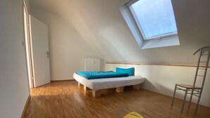 A small, bright room with a slanted ceiling and a skylight, featuring a bed, a chair, and two yellow cushions.
