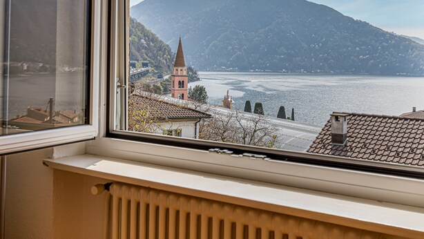 A view from a window overlooking a lake with a mountain in the background and a church with a bell tower. - La Luminosa (Bissone)